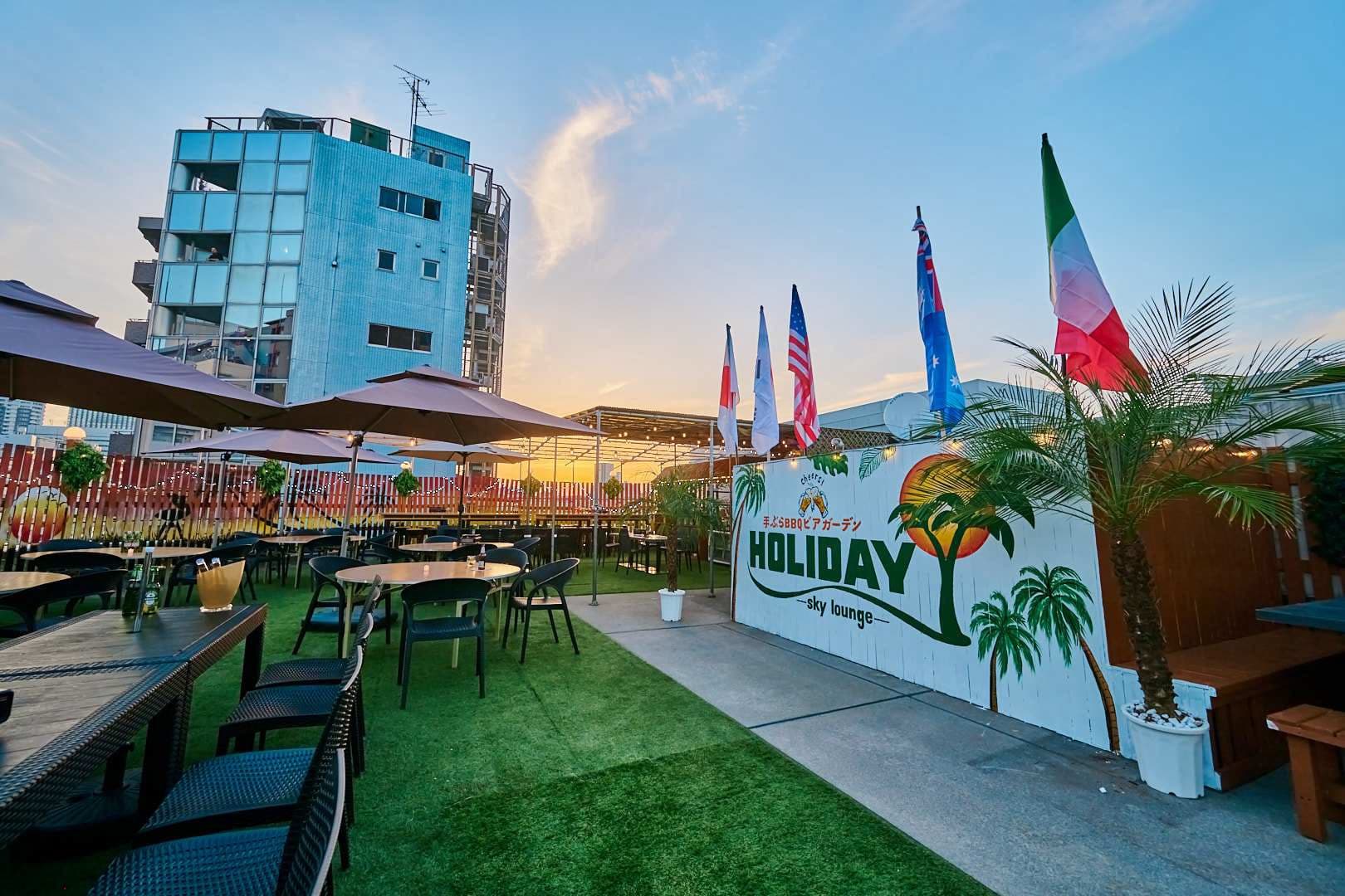 夕暮れの空を背景にヤシの木とHOLIDAYのサインが並ぶテラス席